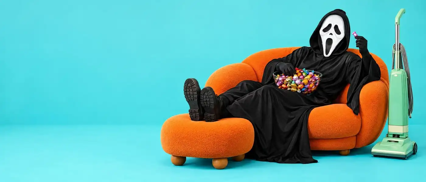 Naamioitunut hahmo lojuu oranssilla sohvalla karkkikulho sylissä, ja imuri seisoo unohdettuna vieressä